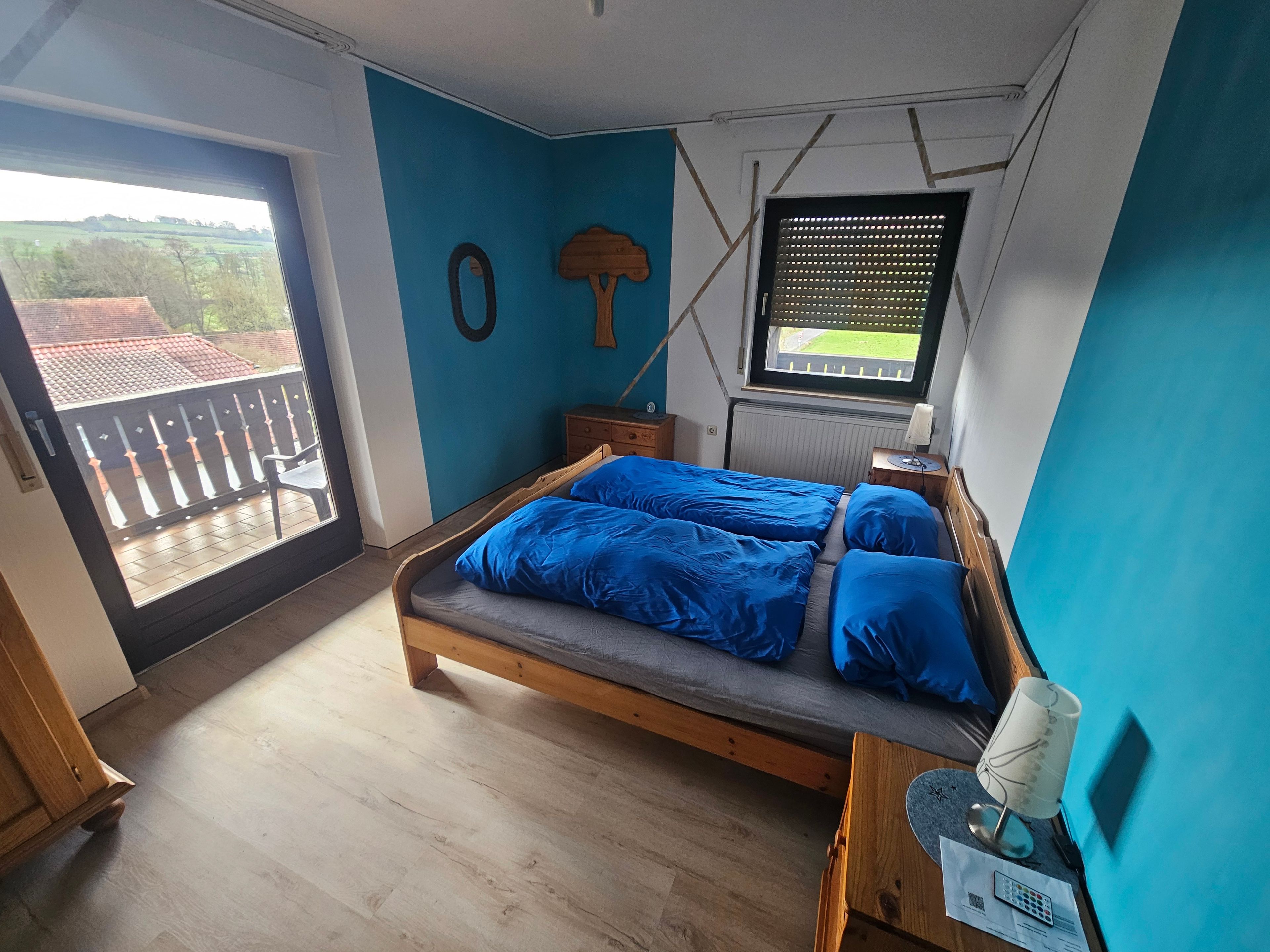 A bedroom with a bed, a window with blinds, a wooden dresser, and a balcony with a view of the countryside.