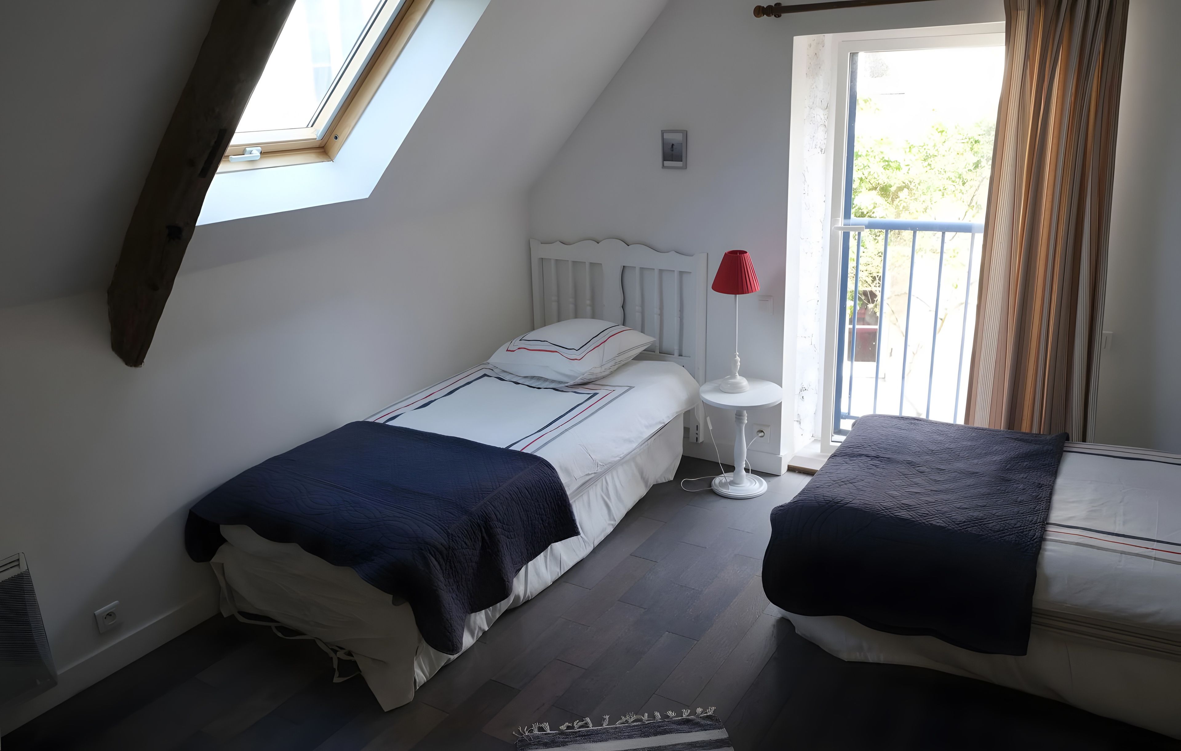 A bedroom with a skylight, a bed with a white headboard, a bedside table with a red lamp, and a window with curtains.
