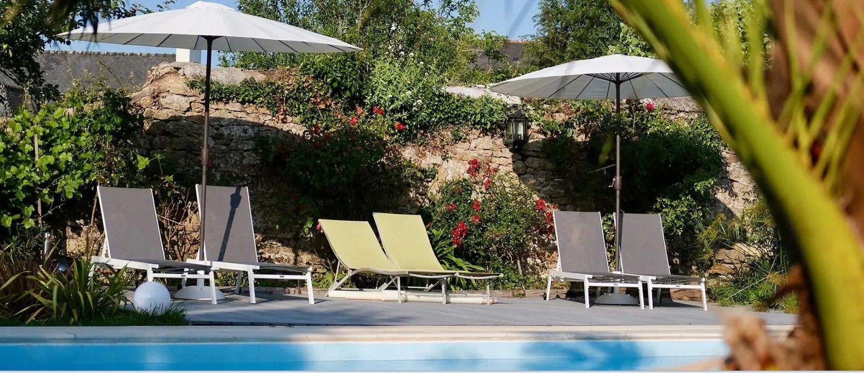 Un espace aménagé au bord de la piscine, avec des chaises longues et des parasols, entouré de verdure et d’un mur en pierre.