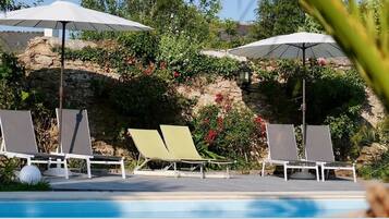Un espace aménagé au bord de la piscine, avec des chaises longues et des parasols, entouré de verdure et d’un mur en pierre.