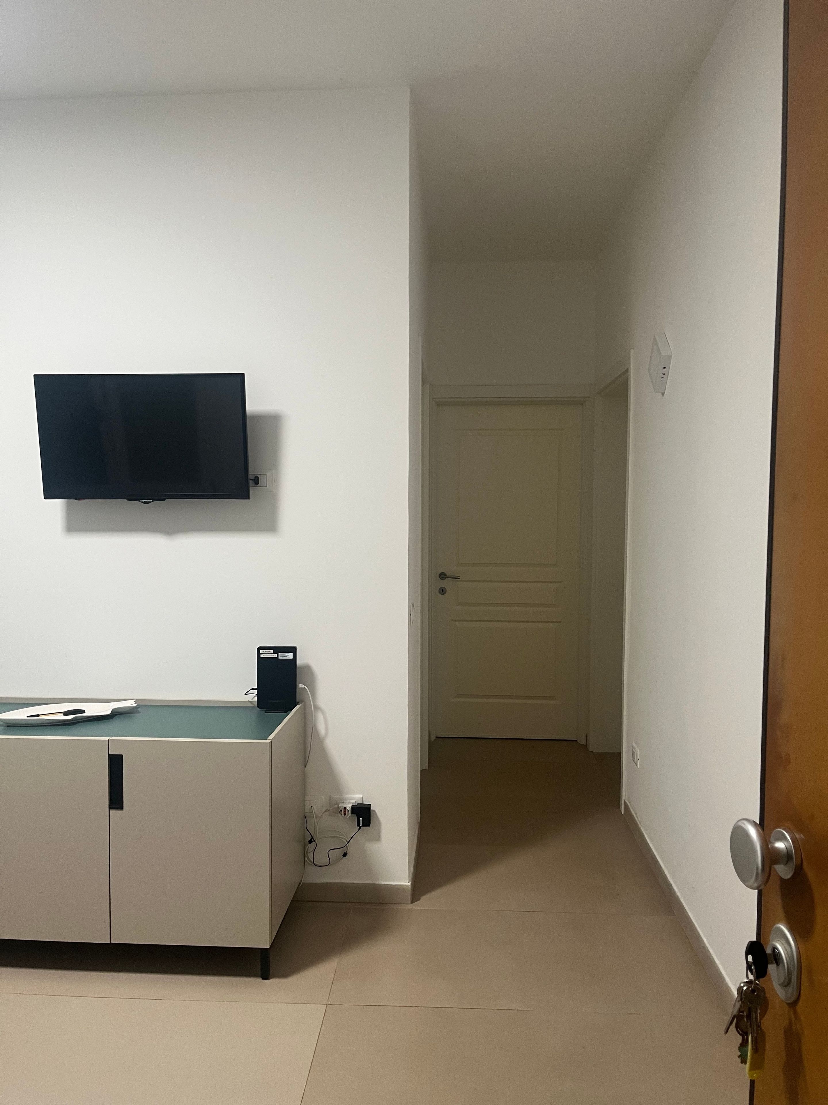 Ein Zimmer mit einem wandmontierten Flachbildfernseher, einem Schreibtisch mit Computer und einer Tür zu einem anderen Raum.