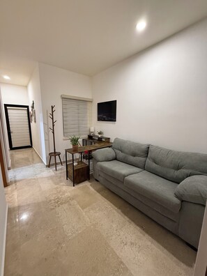 A living room with a grey sofa, a coffee table, a TV mounted on the wall, and a window with blinds. - New Listing (La Paz)
