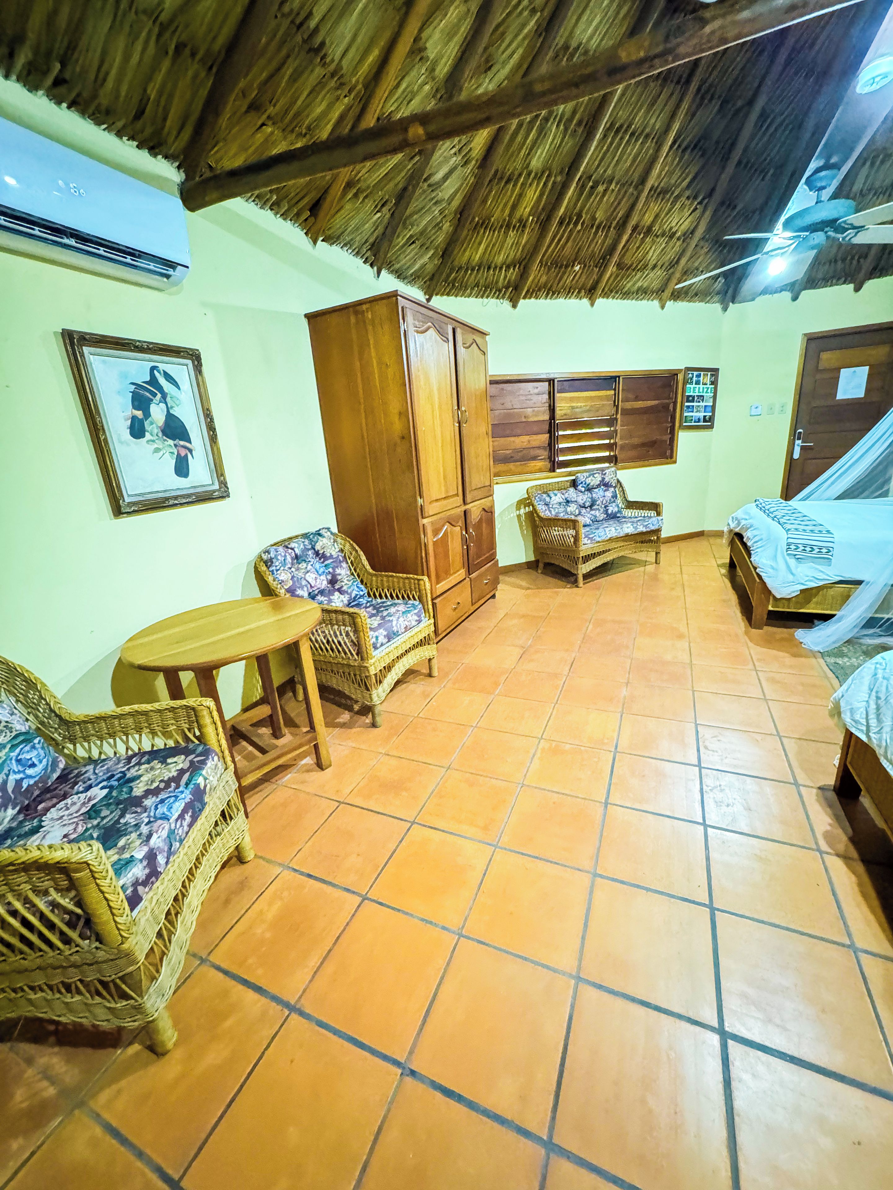 A room with a thatched roof, wicker furniture, a wooden wardrobe, and a bed with mosquito netting.