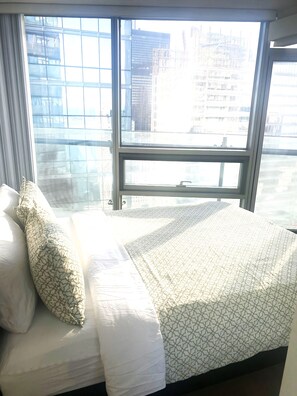 A hotel room with a large window, a bed with white bedding, and a view of city buildings. - Majestic Suites The Union (Queen Bed+Sofa Bed, Up to 3 Guests, Has Crib) (Toronto)