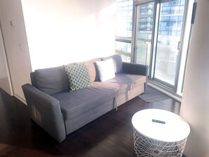 Un moderno salón con un sofá gris, cojines estampados, una mesita redonda blanca y vistas a un edificio a través de amplios ventanales.