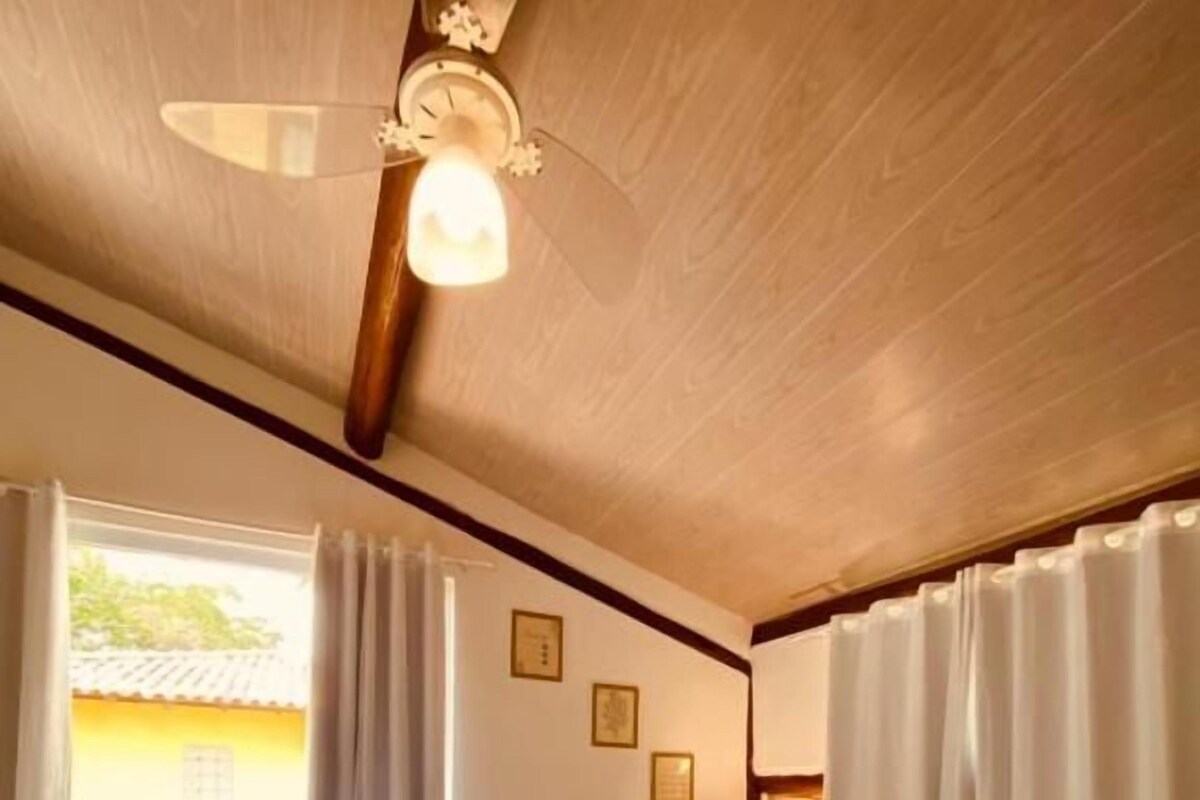 A hotel room with a bed, a ceiling fan, a window with curtains, and a brick wall.