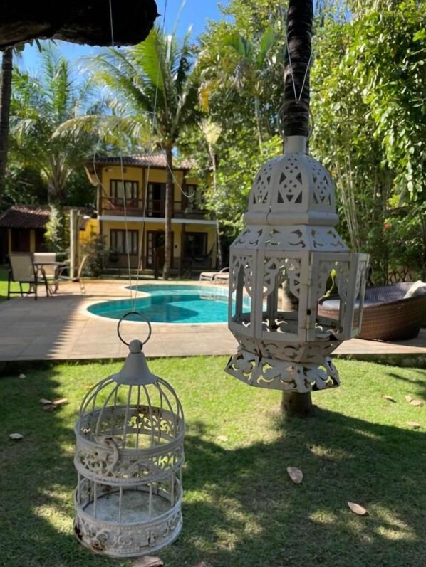 A white birdcage and lantern in a garden with a pool and a house in the background. - Maui Suítes e Chalés (Arraial d'Ajuda)