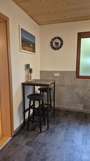 A small room with a wooden ceiling, a framed picture on the wall, a clock with a coffee cup design, a table with two stools, and a window with frosted glass. - Guest House 'Monday's Ferienwohnung' with Wi-Fi (Wehr)