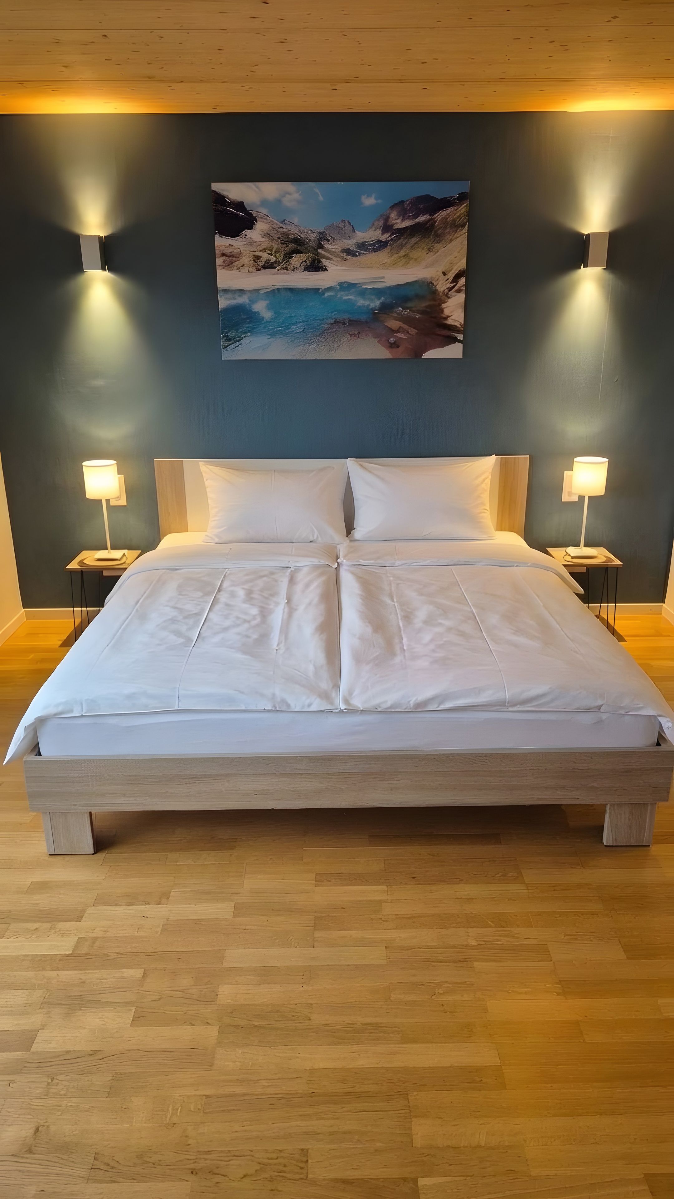 A large bed with a wooden frame and white bedding, flanked by two bedside tables with lamps, in a room with wooden flooring and a painting of a landscape on the wall.