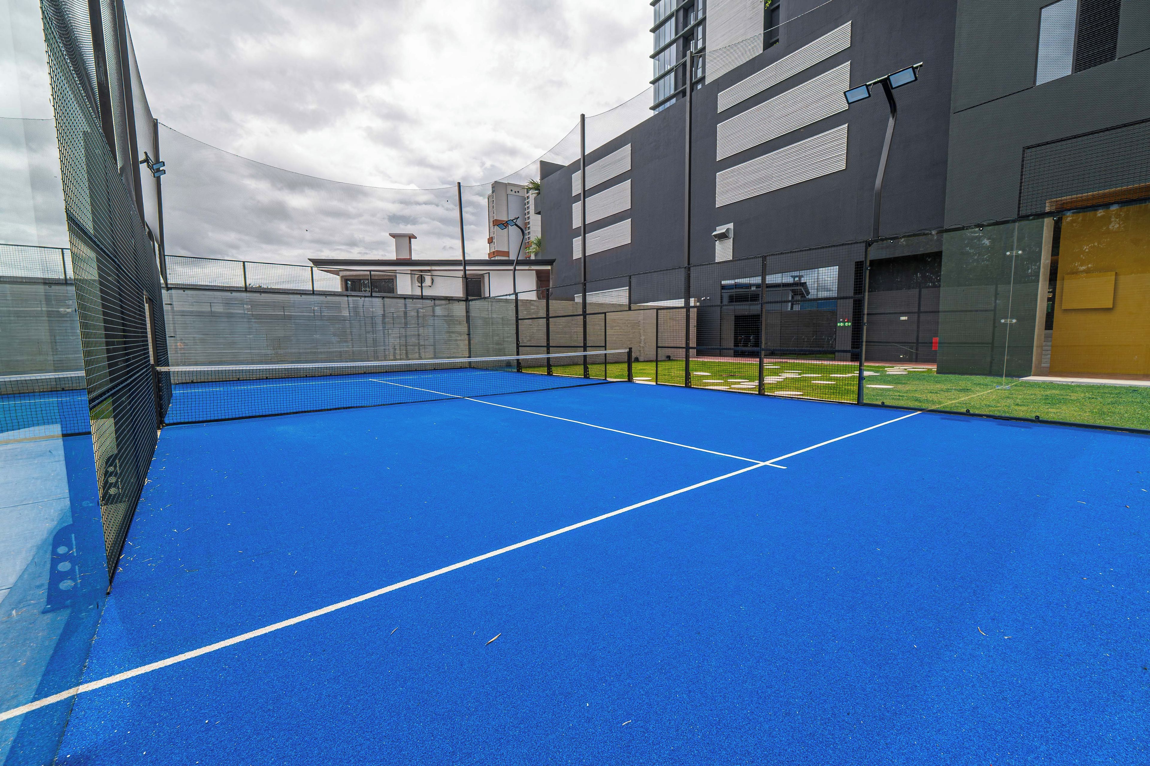 Una cancha de tenis exterior con una red, rodeada por una cerca y edificios cercanos.