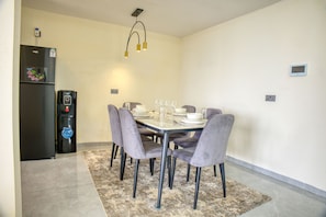 Uma sala de jantar com uma mesa posta para seis pessoas, rodeada por seis cadeiras, uma geladeira preta e um luminário fixado na parede.