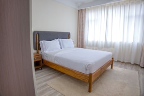 Cama de solteiro com estrutura de madeira e cabeceira cinza, colocada em frente a uma janela com cortinas.