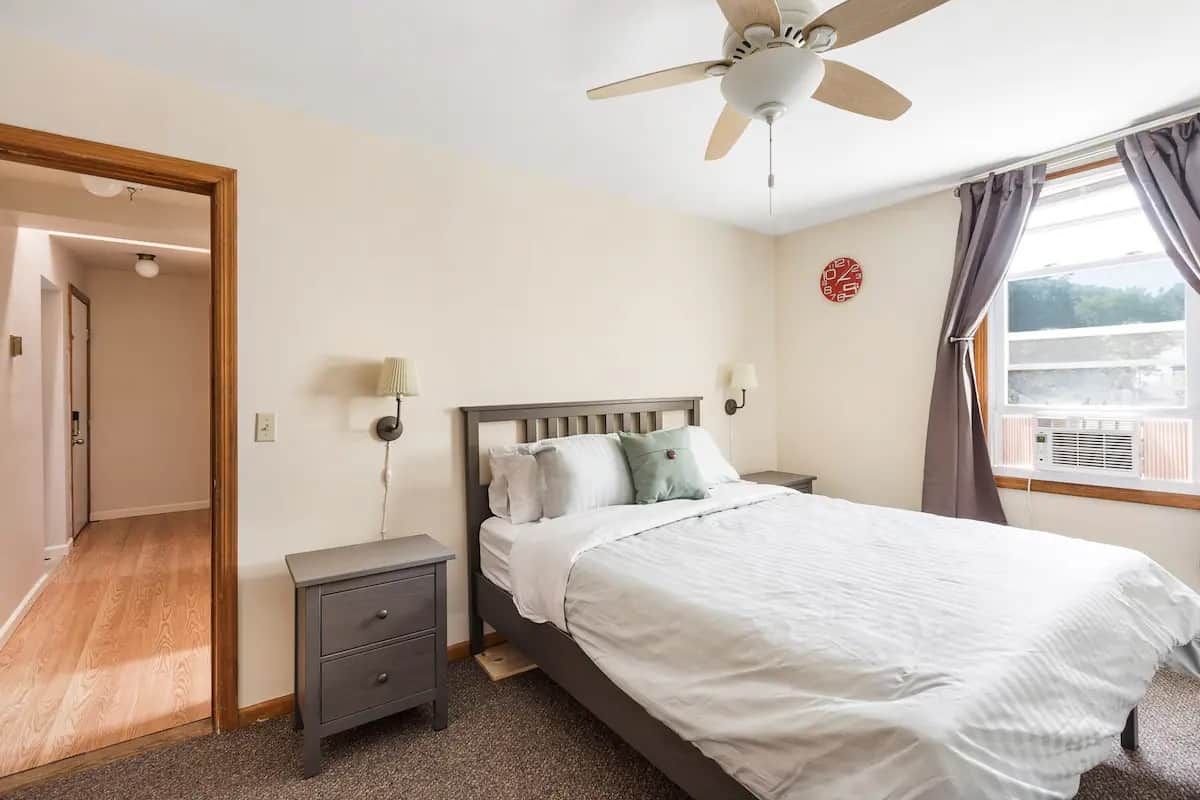 A bedroom with a bed, bedside tables, a ceiling fan, a window with curtains, and a door leading to another room.