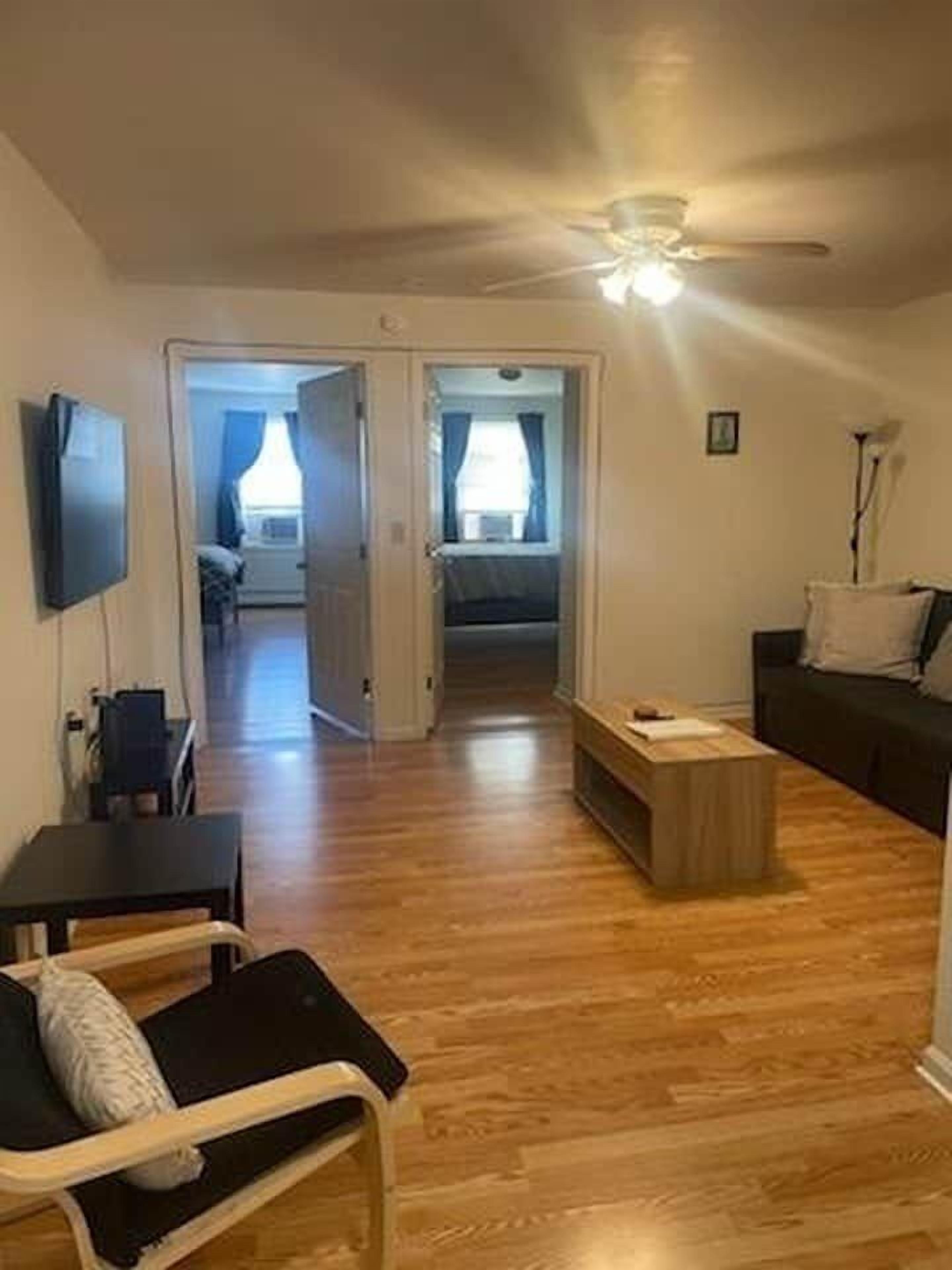 A room with a ceiling fan, a wooden floor, a black sofa, a wooden coffee table, and a TV mounted on the wall.