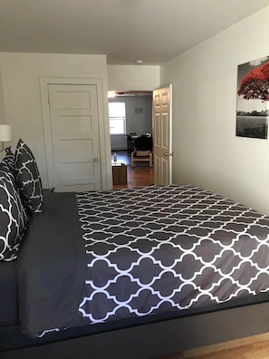 Een slaapkamer met een bed, kussens, een deur en een gang naar een eetruimte.