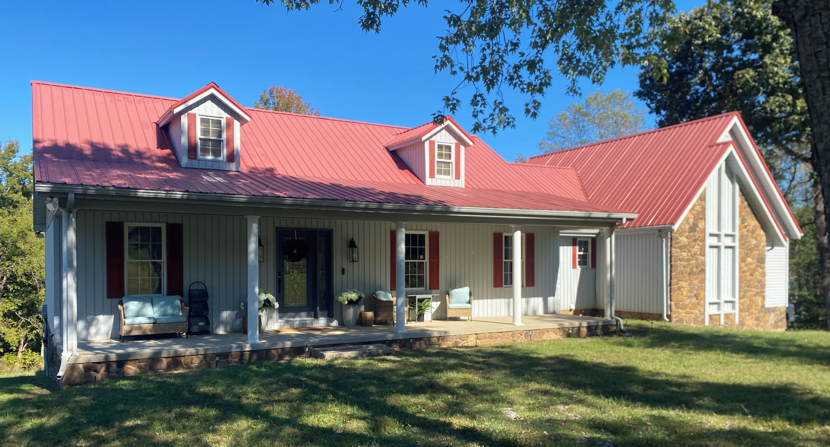 A two-story house with a red roof, a porch with outdoor seating, and a stone foundation.