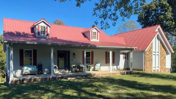 A two-story house with a red roof, a porch with outdoor seating, and a stone foundation.