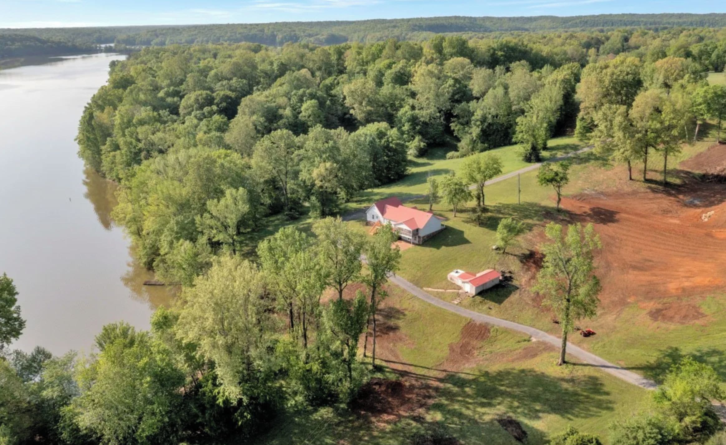 A large house with a red roof, surrounded by a forested area and a body of water.