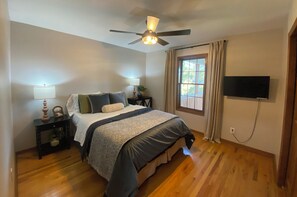 Una camera da letto con un letto, una ventola a soffitto, una finestra con tende e una televisione a schermo piatto montata a parete.
