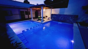 Une piscine avec un patio couvert, un mur en pierre et un petit espace extérieur aménagé pour s’asseoir.