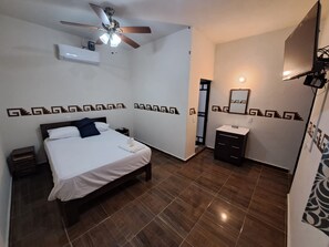 Une chambre d’hôtel comprenant un lit, une table de chevet, un ventilateur de plafond, un appareil de climatisation, une salle de bain avec un lavabo et un miroir, ainsi que des motifs décoratifs sur les murs.