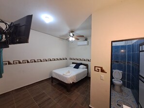Une chambre d’hôtel de taille réduite, comprenant un lit simple, un ventilateur de plafond, un appareil de climatisation et une salle de bain avec des toilettes et une douche.