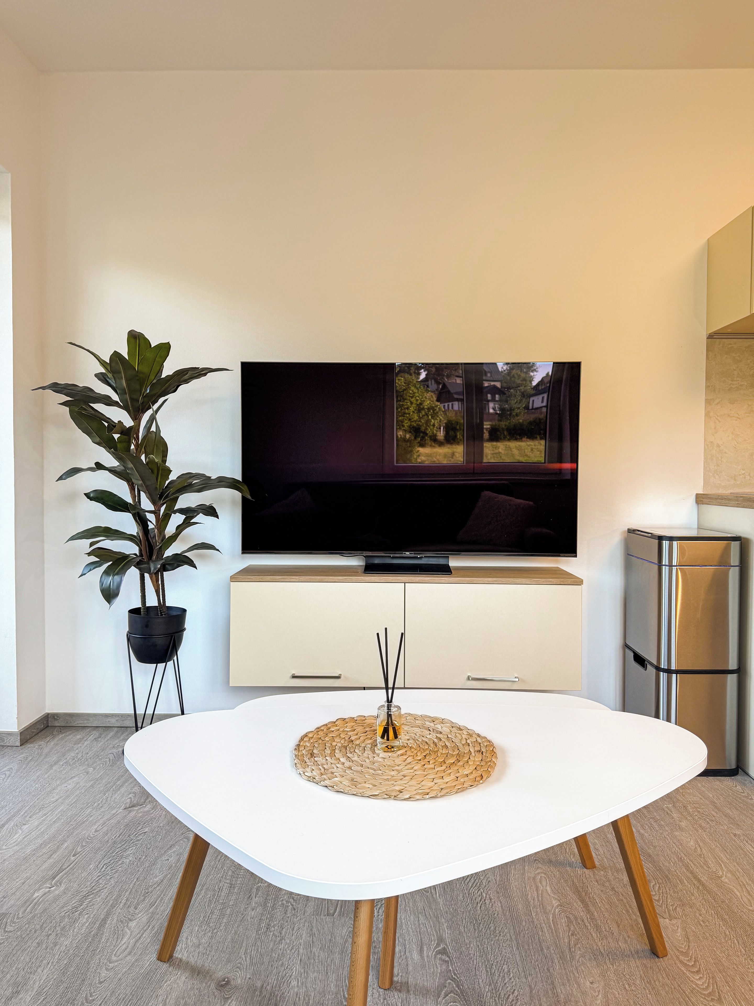 Una sala de estar moderna con un televisor de pantalla plana, una mesa blanca con un mantel tejido, una planta en maceta y una zona de cocina con un refrigerador de acero inoxidable.