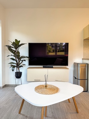 A modern living room with a flat-screen TV, a white table with a woven placemat, a potted plant, and a kitchen area with a stainless steel refrigerator.