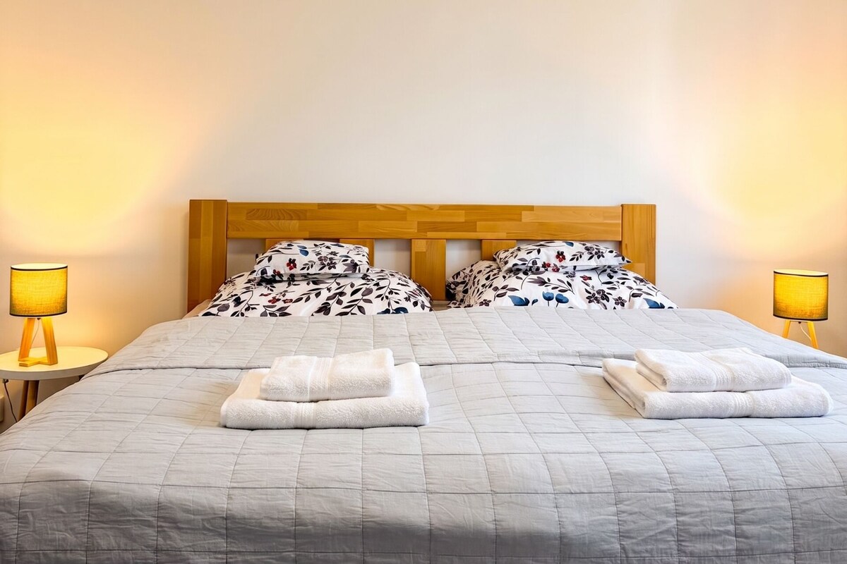 Ein ordentlich bezogenes Bett mit hölzernem Kopfteil, zwei Kissen und gefalteten Handtüchern. Auf beiden Seiten des Bettes stehen je eine Nachttischlampe.