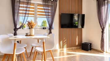 Un comedor moderno con una mesa y sillas blancas, un televisor de pantalla plana montado en un panel de madera y cortinas ligeras.