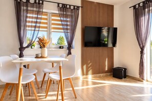 A modern dining area with a white table and chairs, a flat-screen TV mounted on a wooden panel, and sheer curtains.