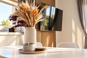Ein modernes Interieur mit einem weißen Tisch, einer Vase mit Trockenpflanzen, einer Kerze und einem Untersetzer. Ein Flachbildfernseher ist an der Wand befestigt.