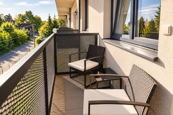 A balcony with two wicker chairs and a small table, overlooking a residential street and greenery.
