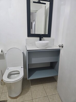 Un baño con inodoro blanco, un lavamanos blanco con grifo negro y un espejo sobre el lavamanos.