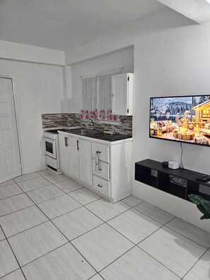 Una cocina compacta con estufa, pileta y gabinetes blancos. Un televisor de pantalla plana está montado en la pared, mostrando un paisaje nevado.