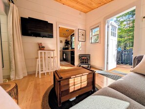 A cozy living room with a wooden coffee table, a sofa, and a TV mounted on the wall. - Tiny House Woodland Retreat • Private Hot Tub • Sugarbush (Warren)