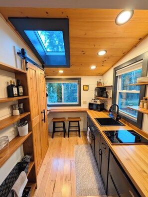 A compact kitchen with wooden cabinets, a skylight, and a view of the outdoors. - Tiny House Woodland Retreat • Private Hot Tub • Sugarbush (Warren)