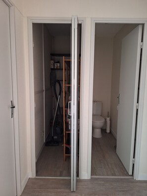 Une pièce avec un plancher en bois, une porte blanche, des toilettes blanches et une étagère contenant des produits de nettoyage.