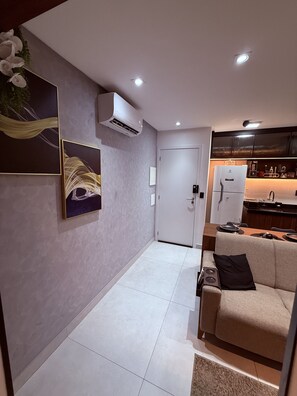 Una sala de estar moderna con un sofá, una mesa de centro y un televisor. Hay una zona de cocina con un refrigerador y un lavamanos. El piso es de baldosas y hay una puerta que da a otra habitación.