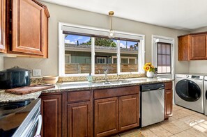 A kitchen with wooden cabinets, a granite countertop, a built-in dishwasher, and a window with a view of a house. - Spacious Furnished 5BR Denver Home, Pet-Friendly, Fenced Yard & Garage (Denver)