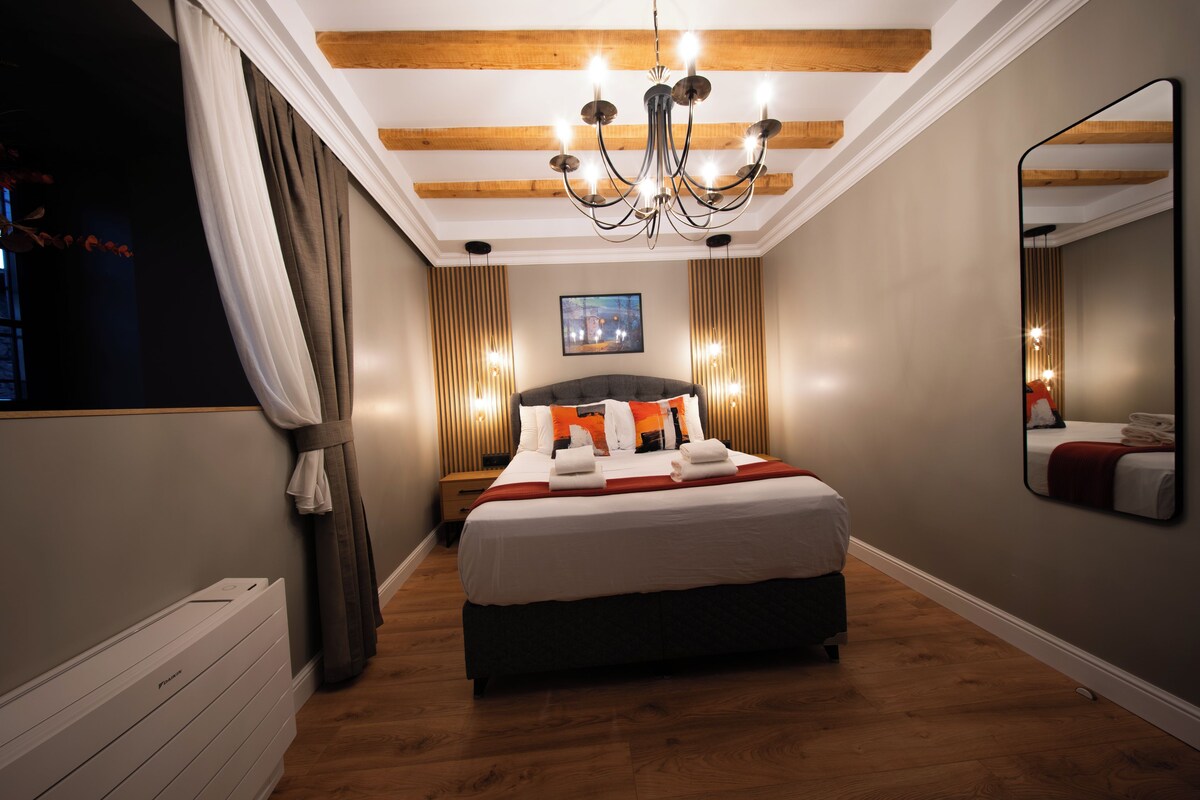 Quarto de hotel com uma cama grande, vigas de madeira no teto, um candelabro e um espelho na parede.