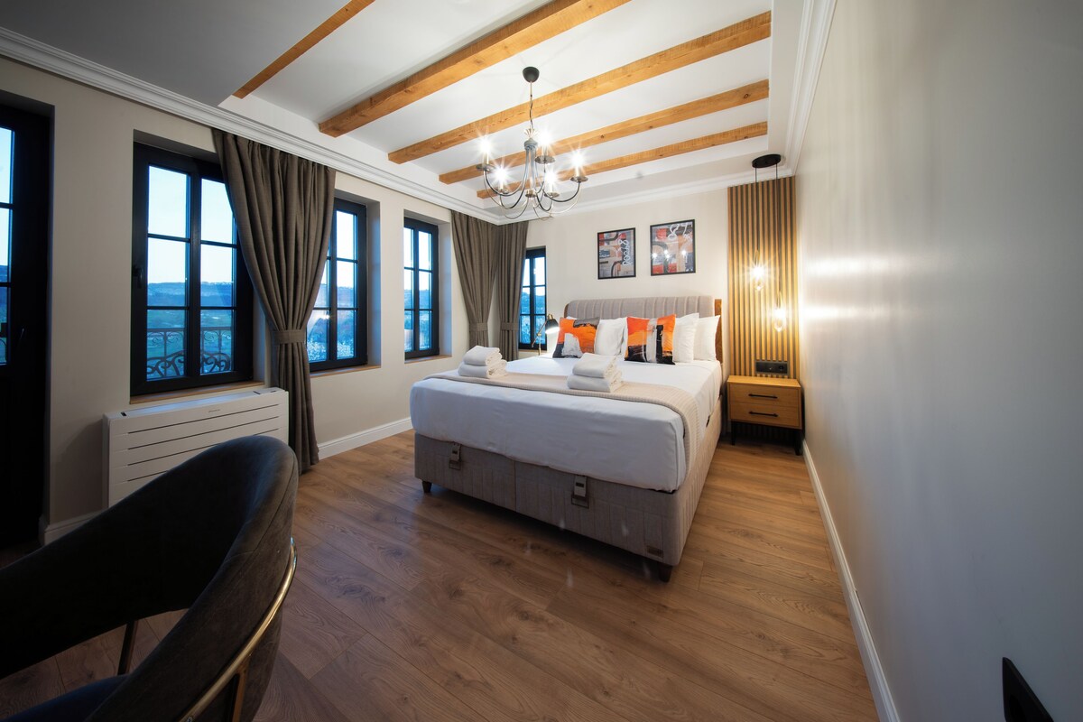 Quarto com uma cama grande, uma escrivaninha e uma cadeira. Há janelas com cortinas, um candelabro e obras de arte na parede.