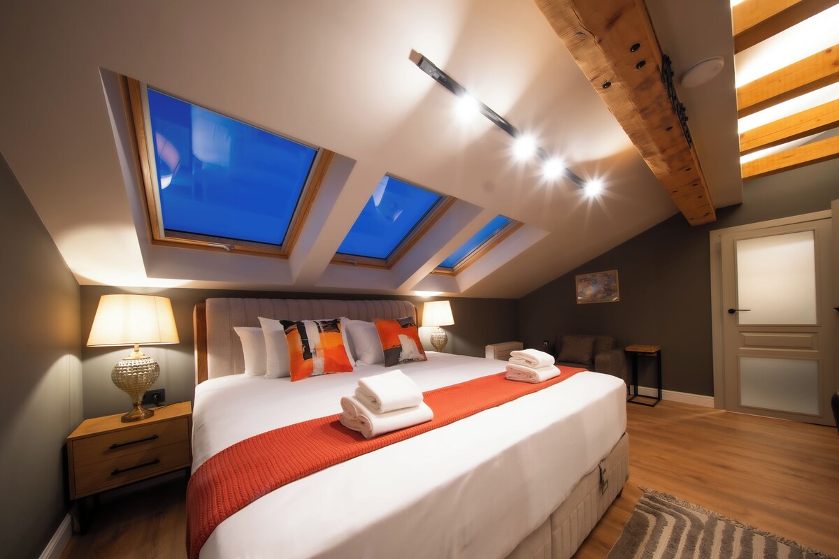 Quarto moderno com cama grande, claraboias, mesa de cabeceira e uma porta.