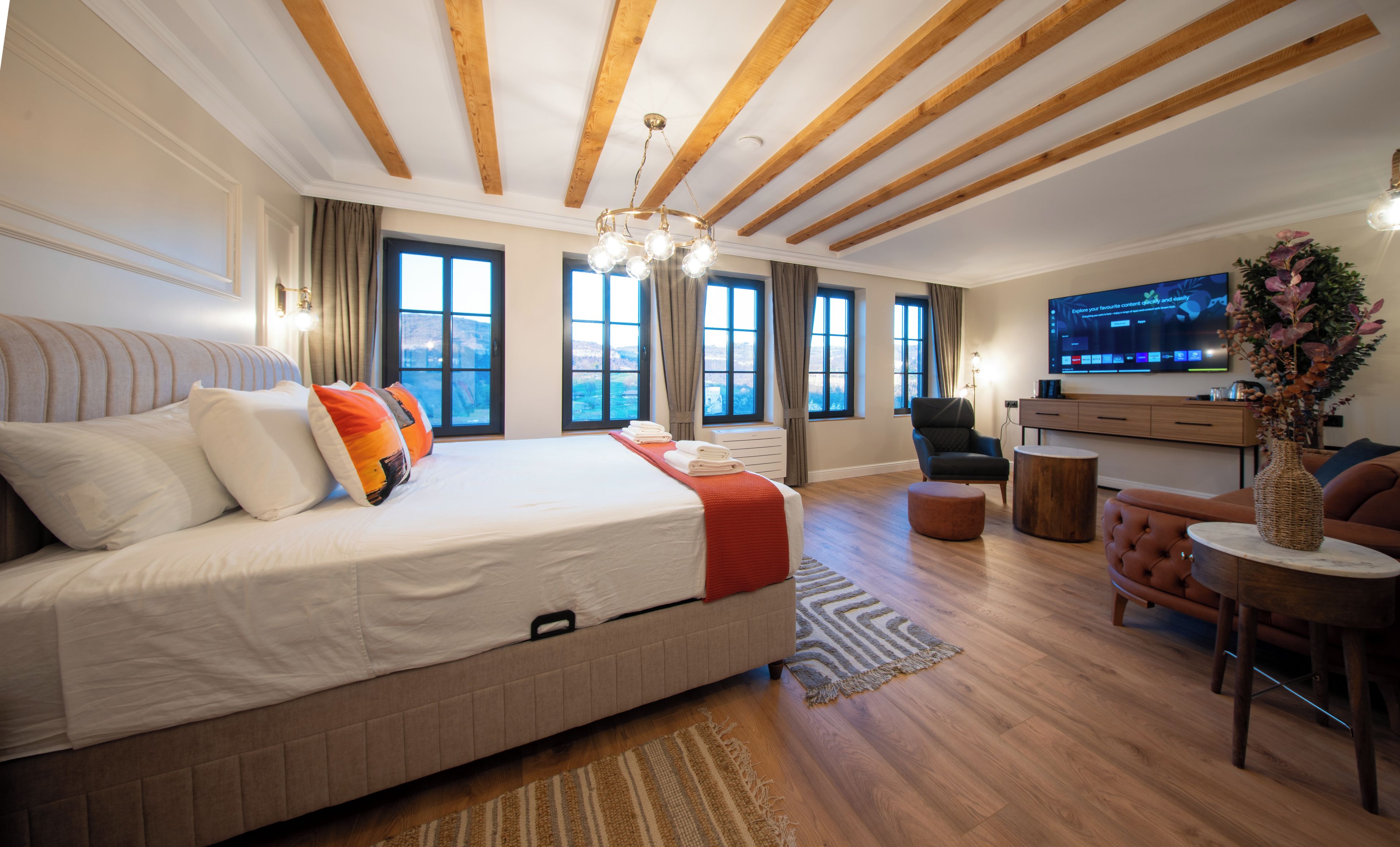 Ein modernes Hotelzimmer mit einem großen Bett, einer Sitzecke inklusive Sofa und Sessel, einem Fernseher und Blick ins Freie.