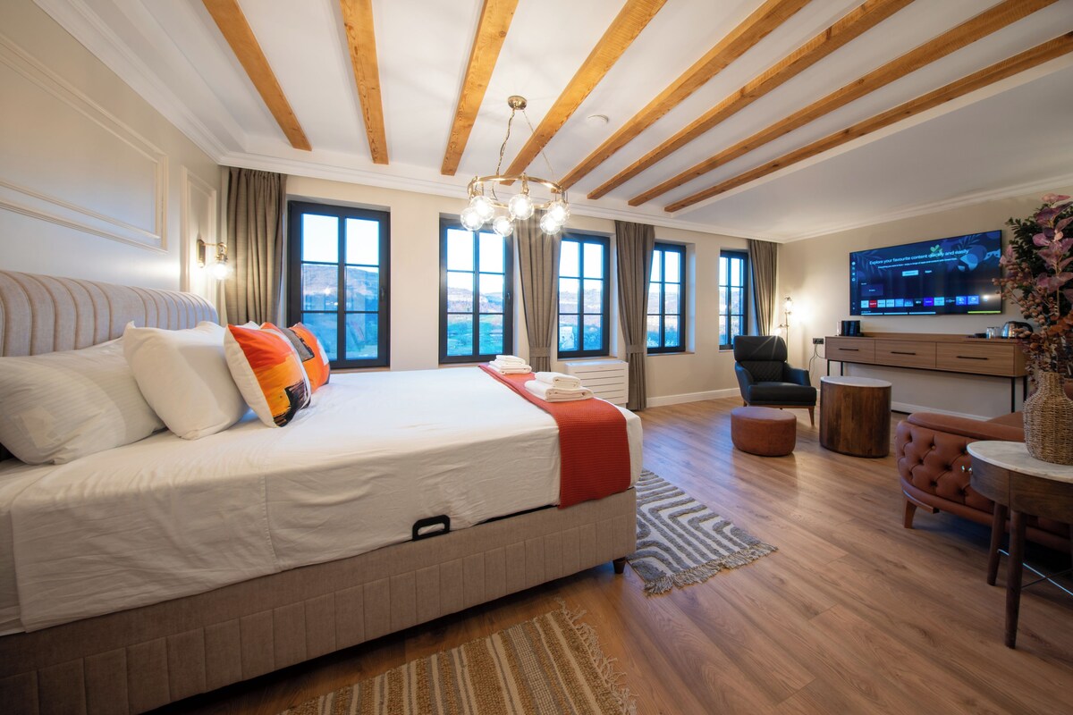 Quarto de hotel moderno com uma cama grande, área de estar com sofá e poltrona, televisão e vista para o exterior.