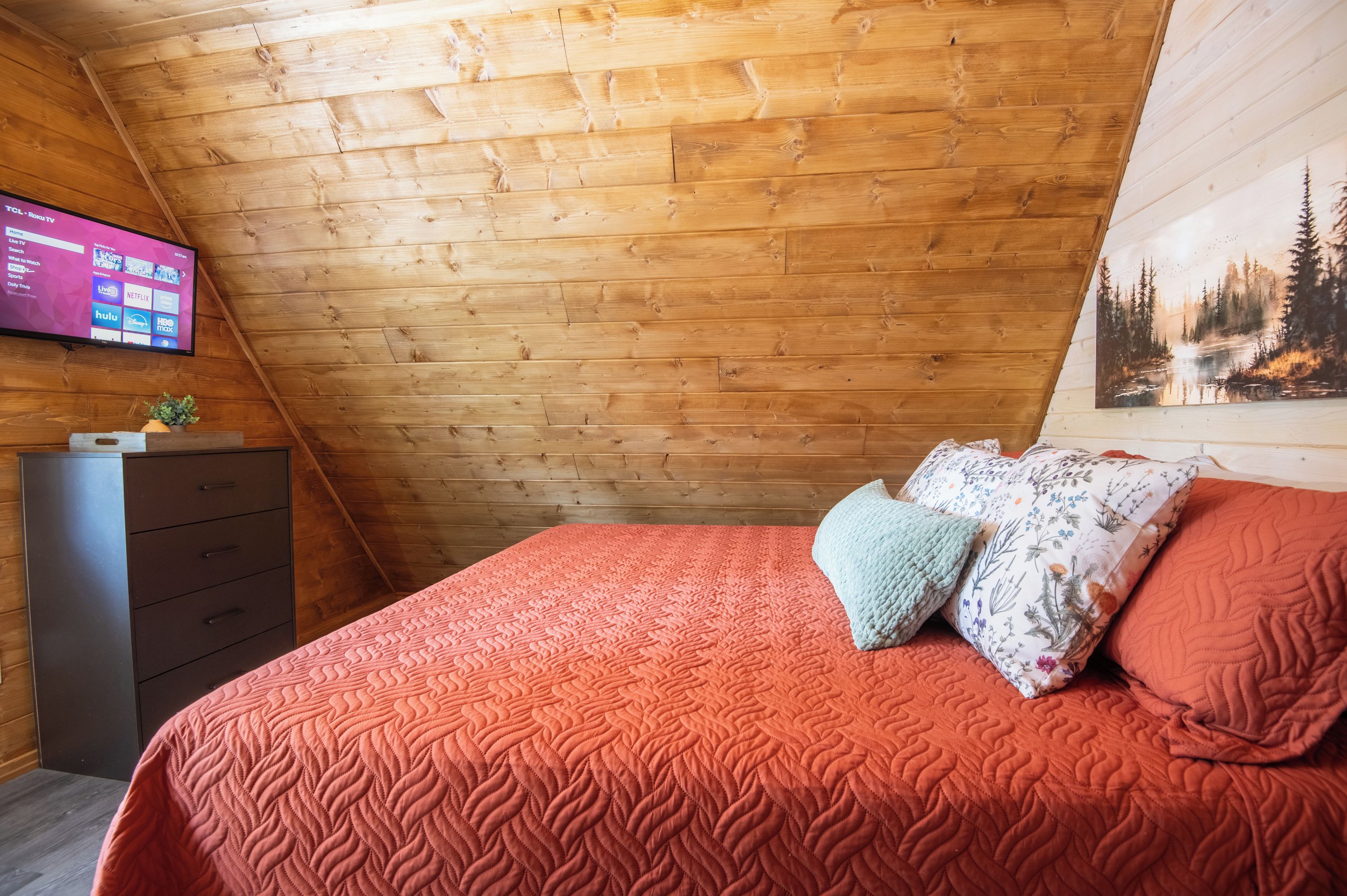 Ein Schlafzimmer mit schrägem Dach, einem Bett mit roter Steppdecke, einem Schminktisch und einem an der Wand befestigten Flachbildfernseher, der einen Fernsehprogramm-Guide anzeigt.