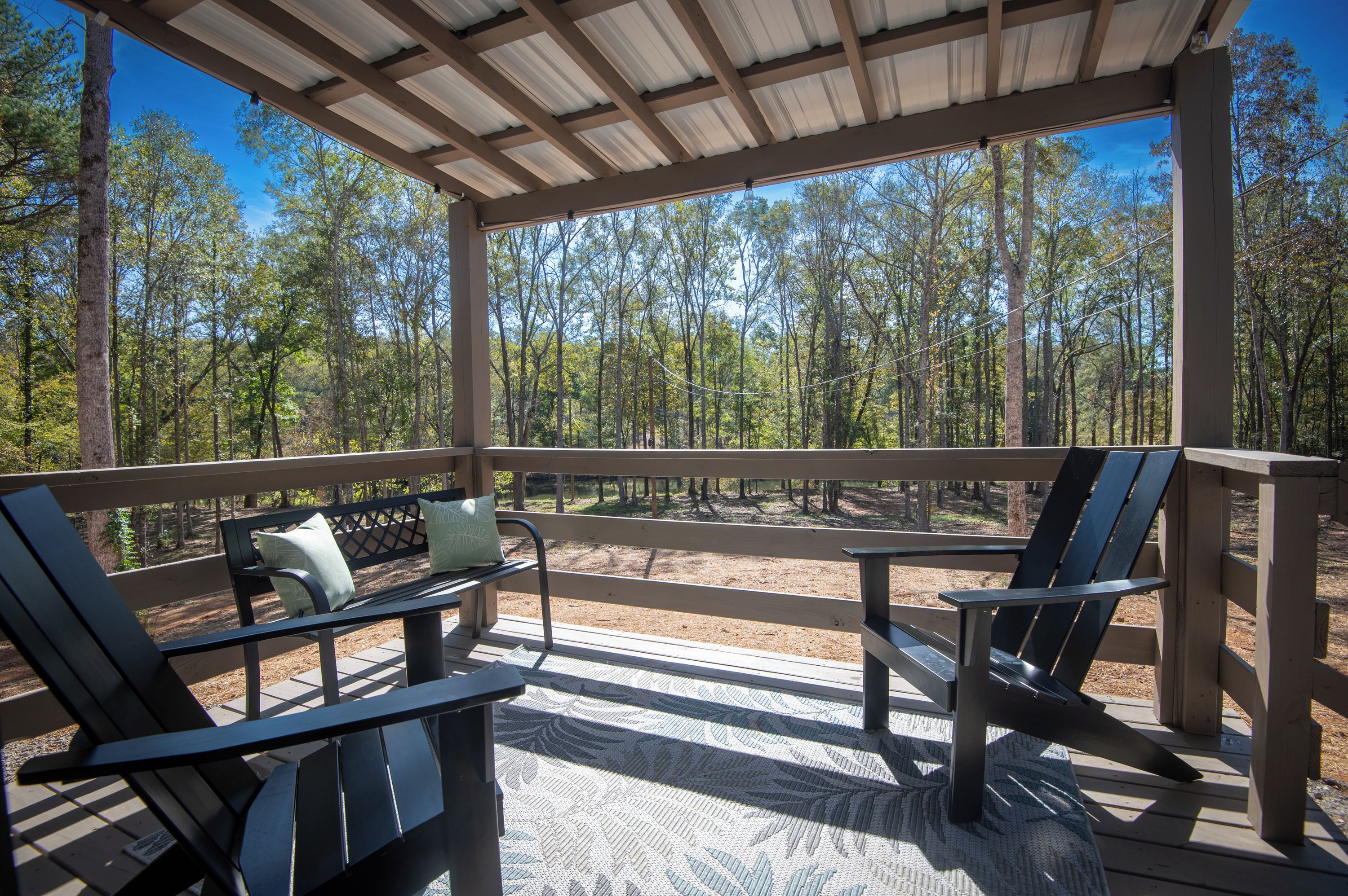 A covered outdoor seating area with two chairs and a table, surrounded by a wooden railing, overlooking a forested area.