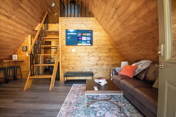 A cozy loft-style room with a wooden interior, a staircase, a mounted TV, a leather sofa, and a patterned rug.
