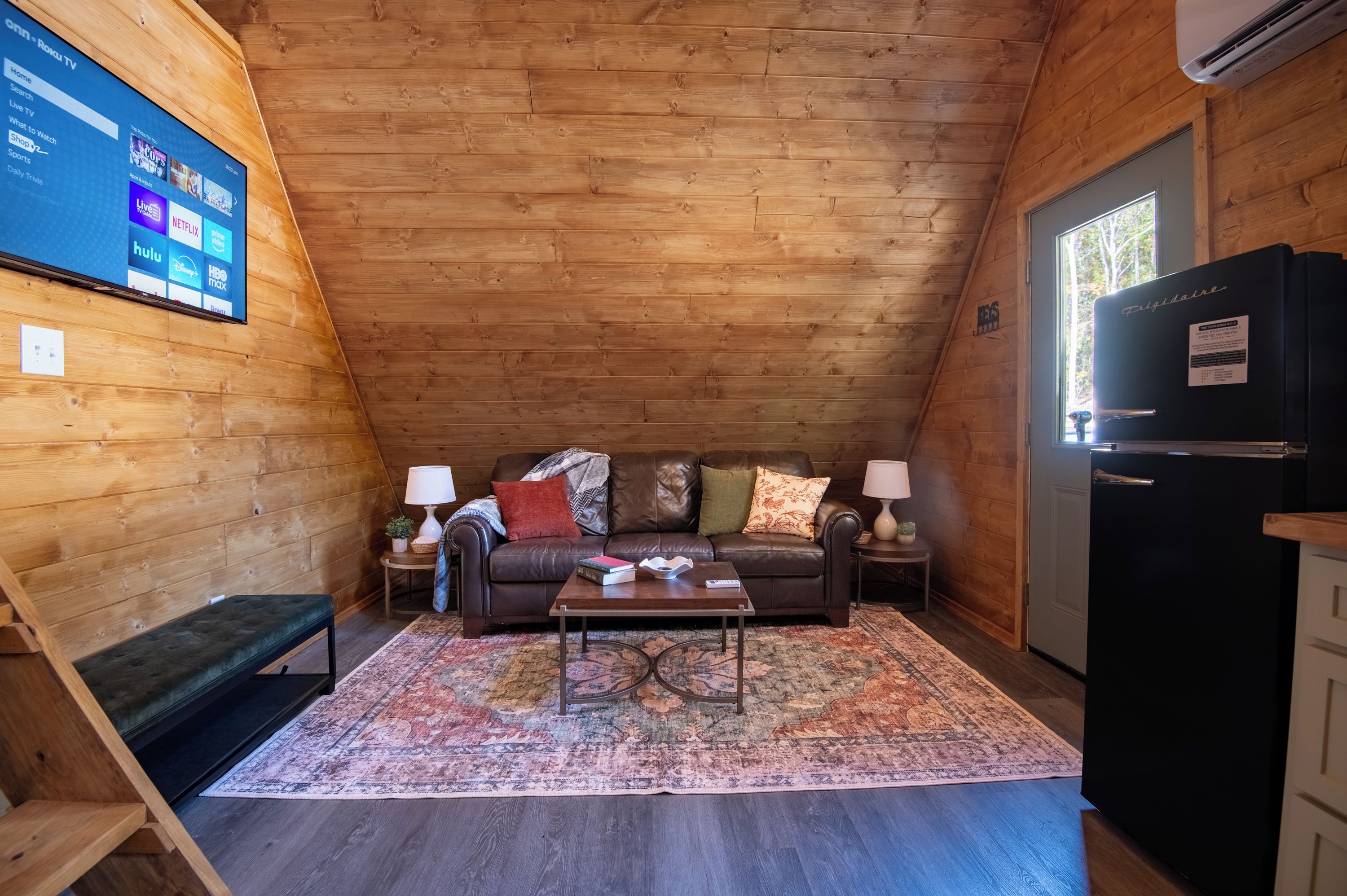 Ein gemütliches Zimmer mit Holzbalkendecke, einem schwarzen Ledersofa, einem Couchtisch, einem gemusterten Teppich und einem schwarzen Kühlschrank.