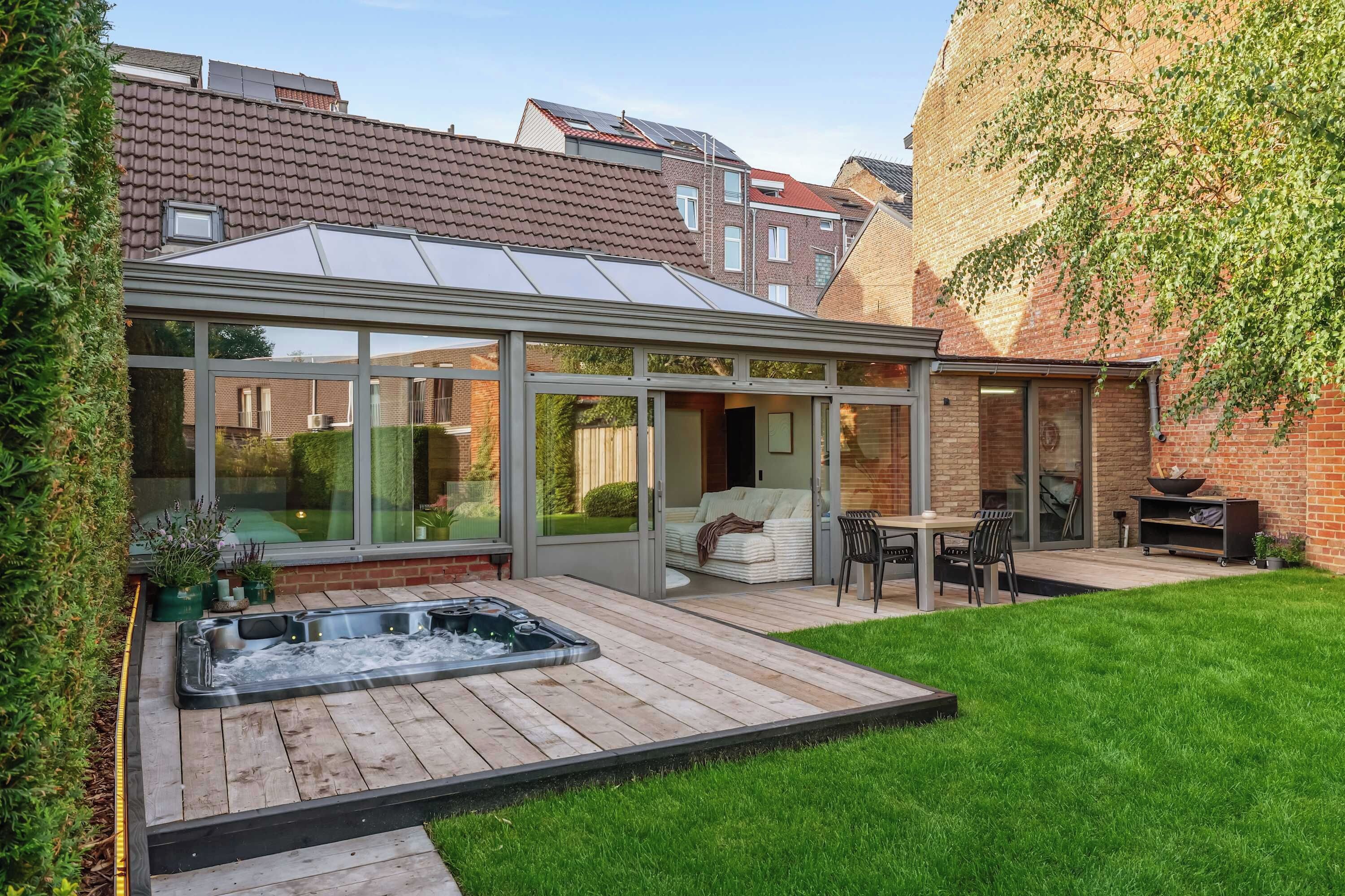 A modern outdoor patio with a hot tub, seating area, and a glass-enclosed space with a view of the garden.